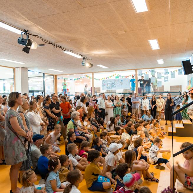 Kinder und Eltern in der Aula stehen und sitzen am Boden